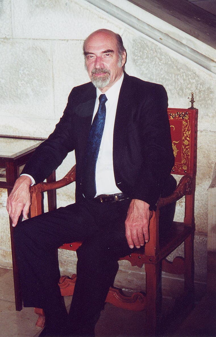 Bernhard Banaschewski in a dark suit, seated in a carved wooden chair against a stone wall